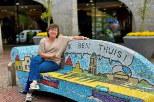 Smiling woman with glasses sits on a colorful mosaic bench outdoors near a modern building and planters with yellow flowers.